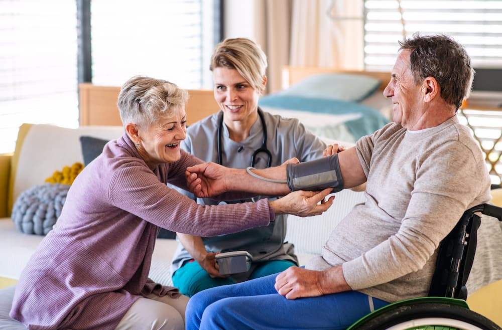 A caregiver checking an older man's blood pressure while his wife holds his hand